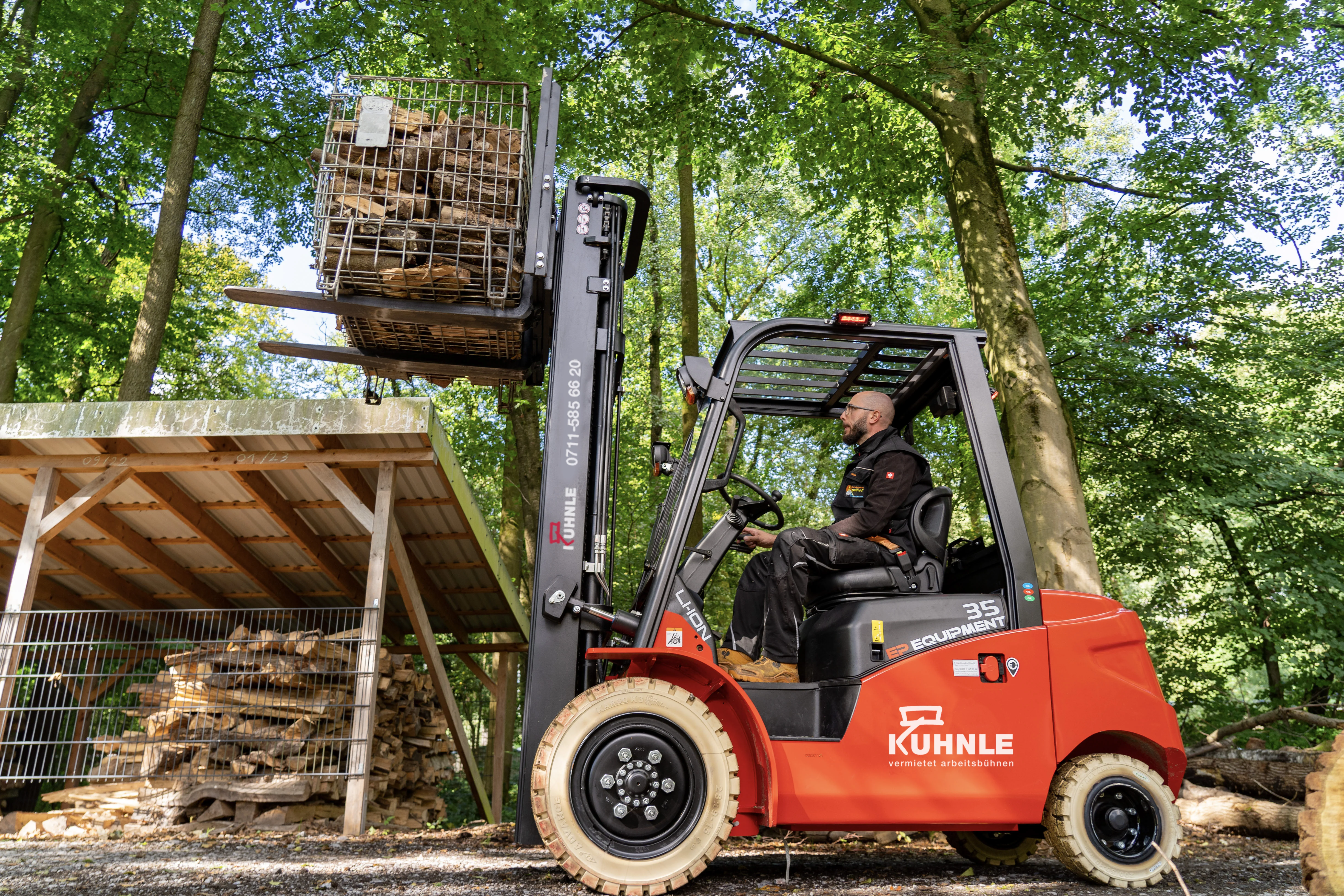 Seitlich fotografierter Stapler mit Person darin, geladen mit Holz; links überdachtes Holzlager, Wald im Hintergrund.